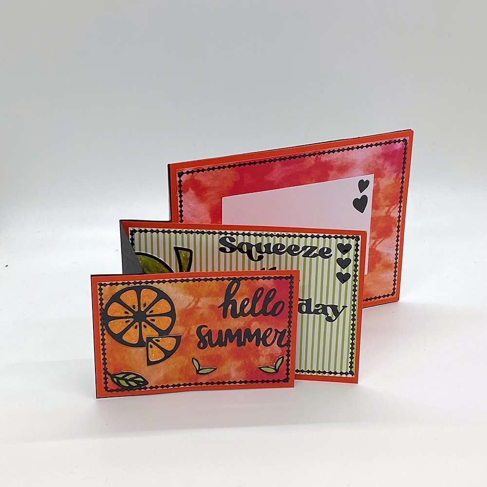 Three tiered orange stand-up card with a small orange citrus slice panel in front, a green striped panel behind it and a red and pink watercolor panel at the back, with black outlines and heart accents. Text reads: "hello summer" - Grammy's Grams Handmade Greeting Cards